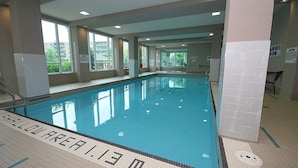 Una piscina interior con señalización clara, incluyendo un letrero que indica 'SHALLOW AREA', rodeada de ventanas y paredes de azulejos.