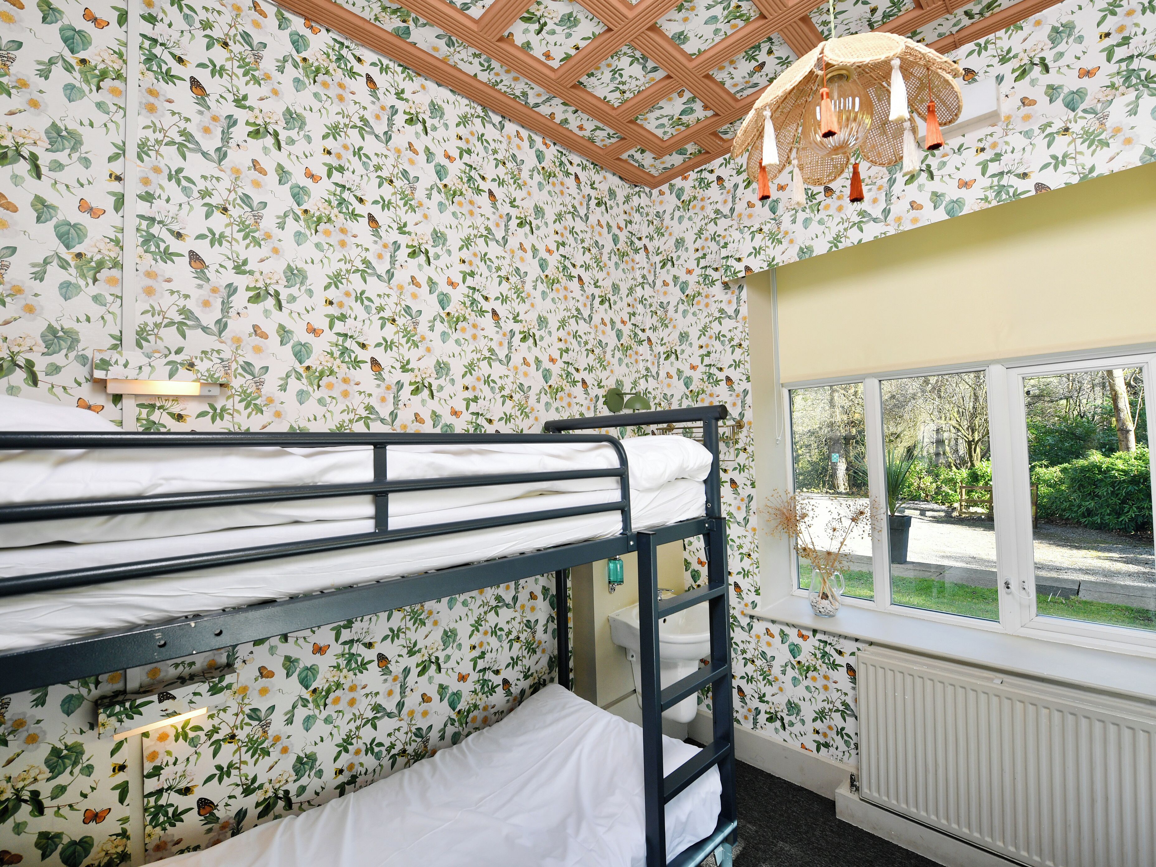 Une chambre avec un papier peint à motifs floraux, un lit superposé, une fenêtre donnant sur la verdure et un ventilateur de plafond.