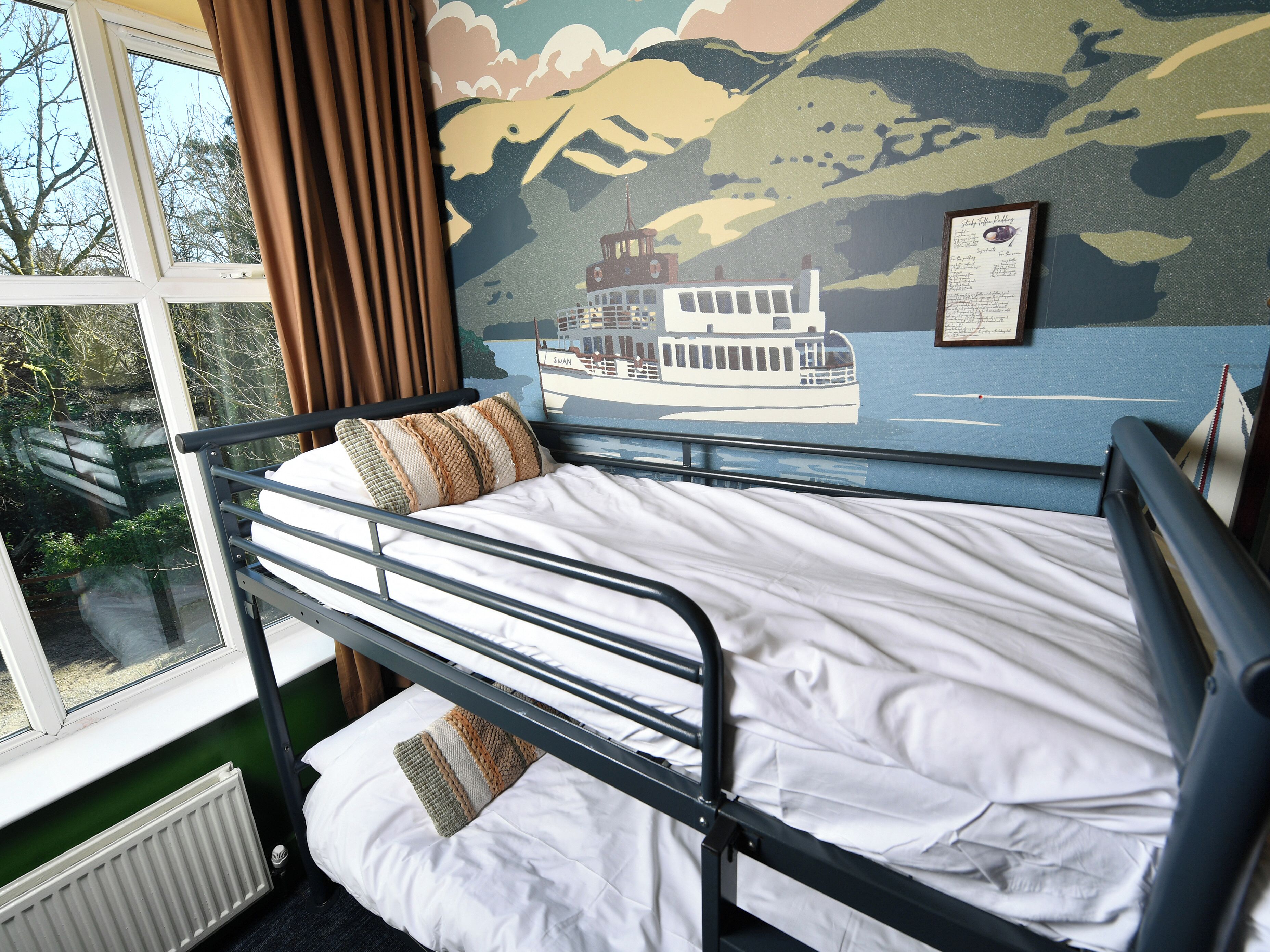 A bunk bed with a white mattress and a pillow, a window with curtains, and a mural of a boat on a river.