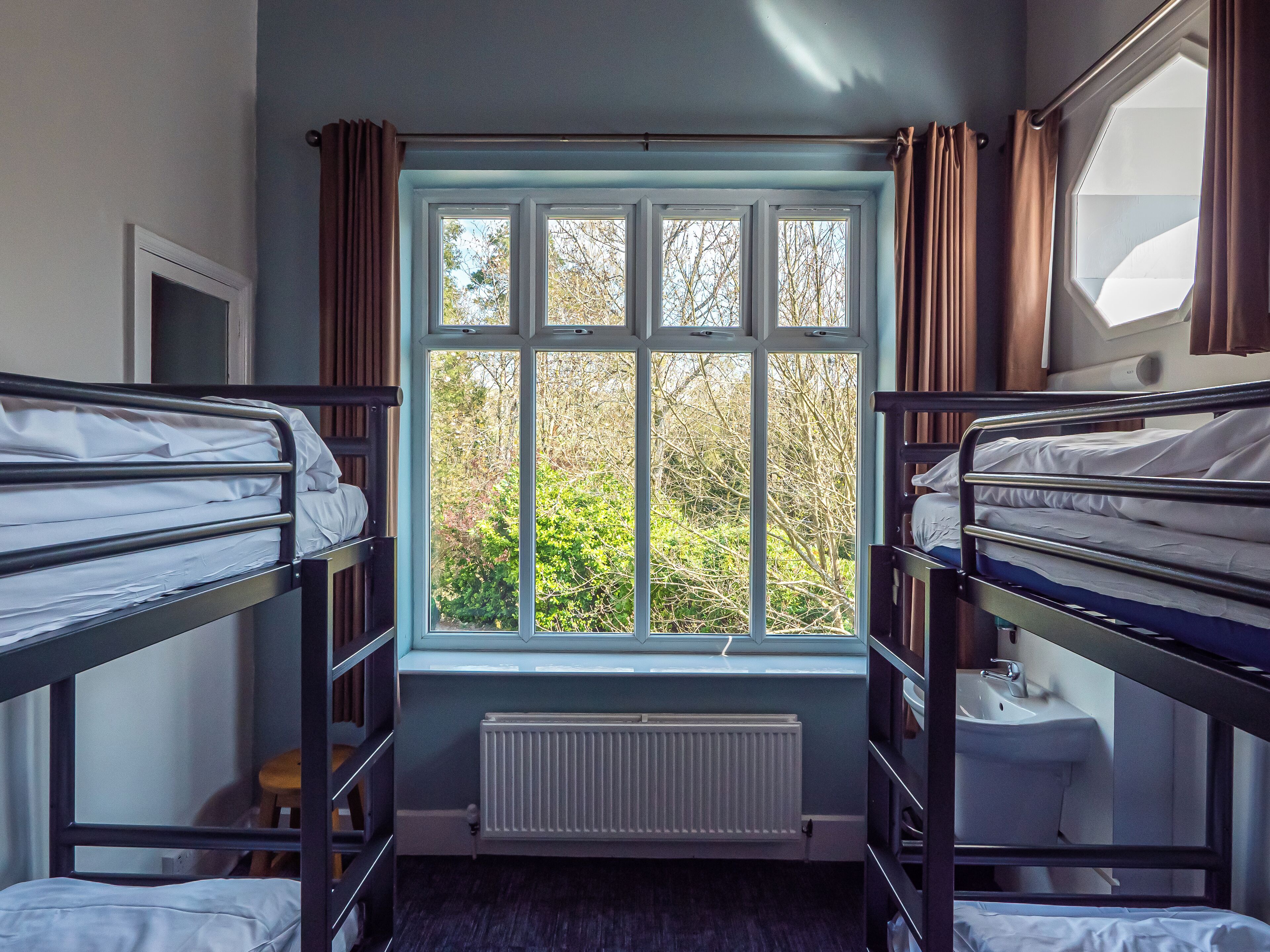 A dormitory room with bunk beds, a window with curtains, a radiator, and a sink.