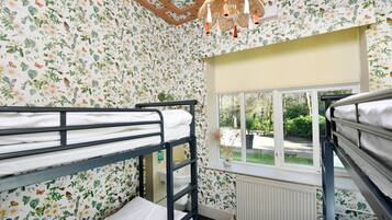 Una habitación con dos literas, papel tapiz floral, una ventana con vista a follaje y una lámpara colgante.