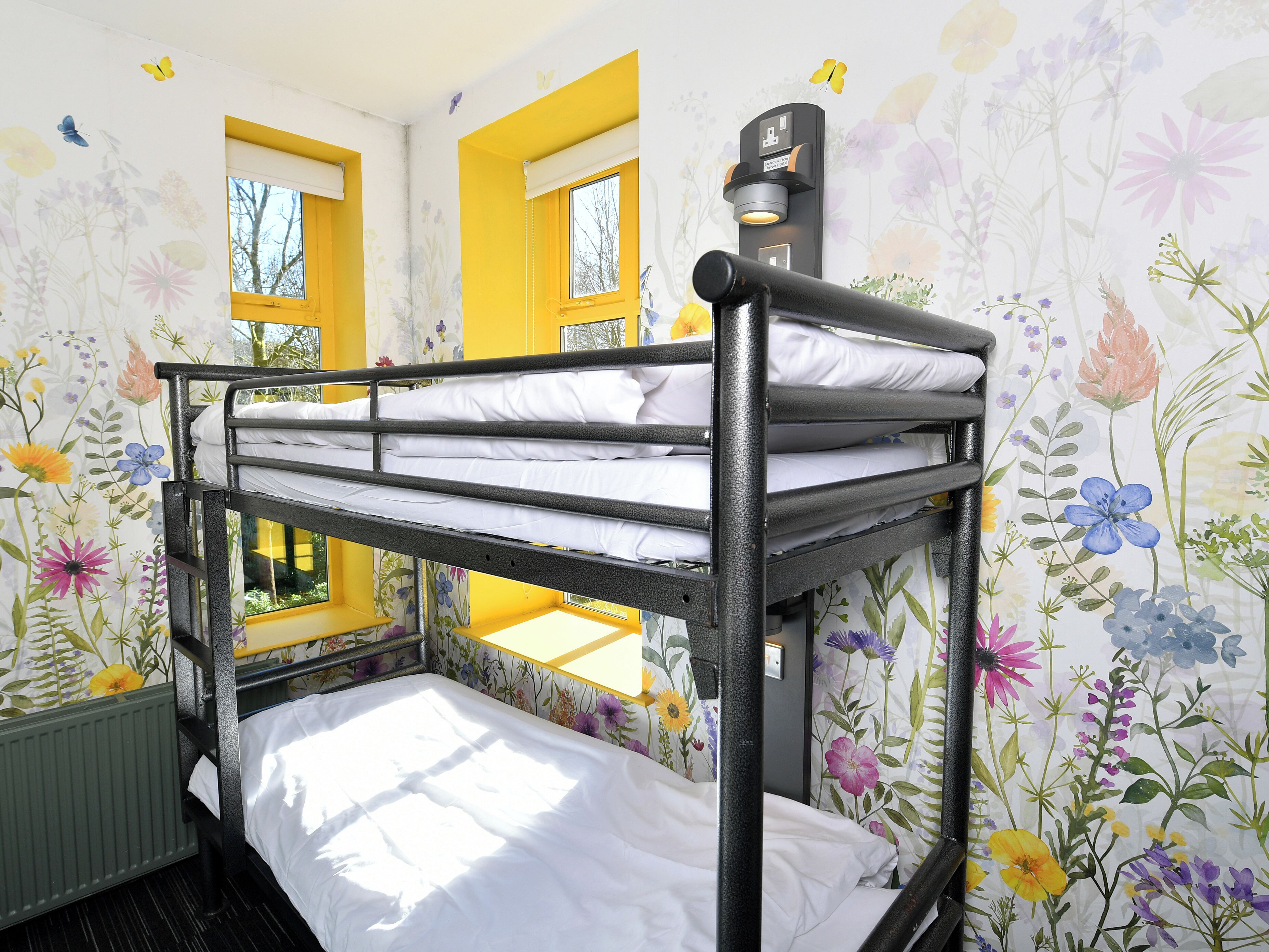 Un lit superposé dans une chambre dont le papier peint est à motifs floraux, avec deux fenêtres et un luminaire fixé au mur.