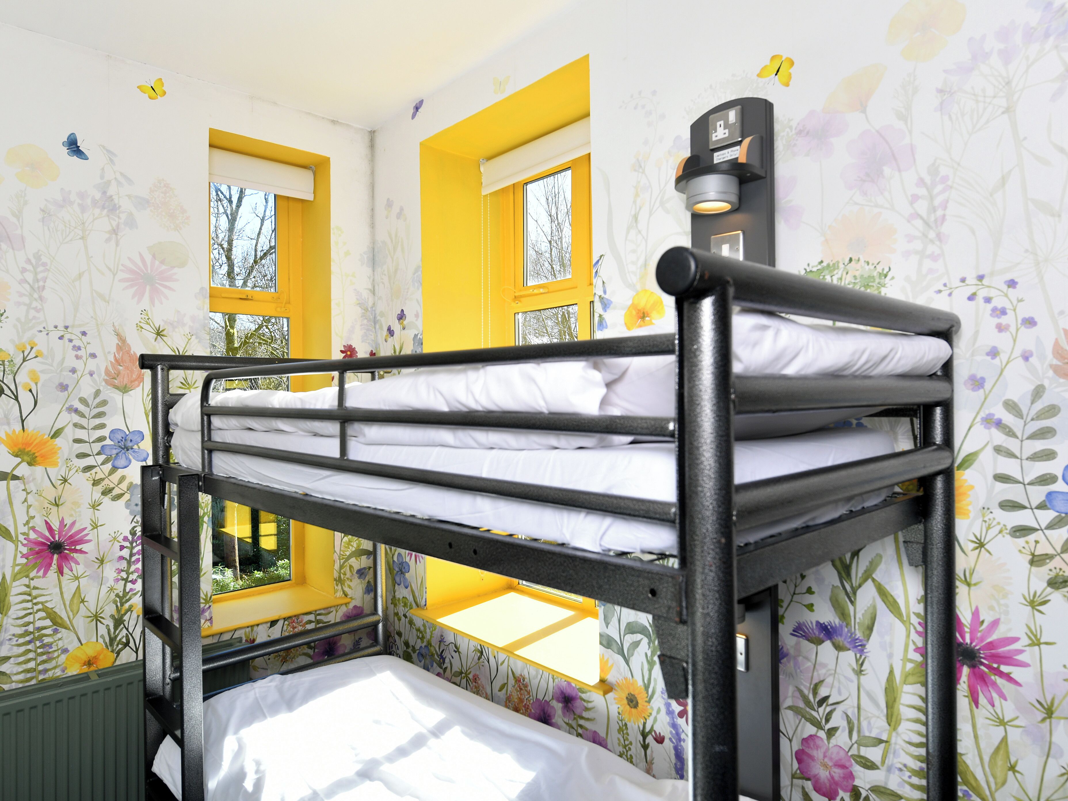 Dormitorio con literas, papel tapiz floral, detalles en amarillo y ventana con vistas.