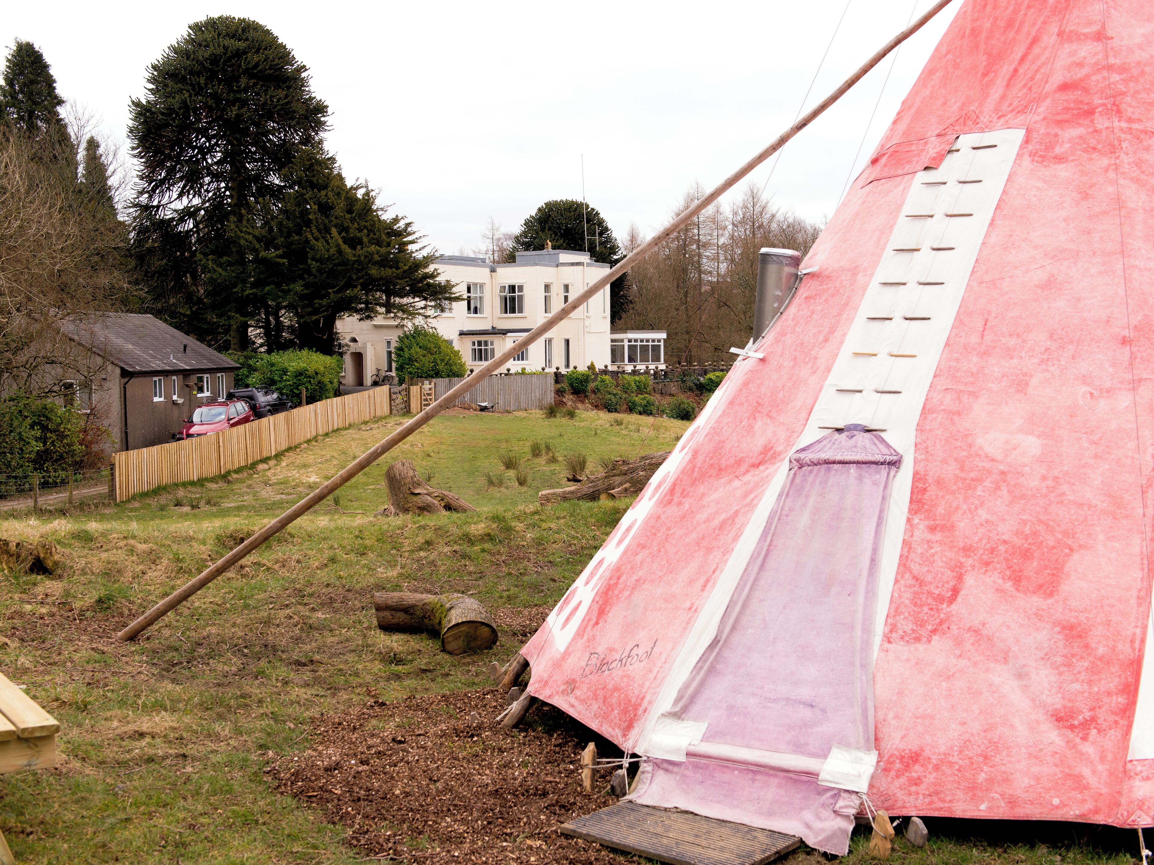 Uma tenda rosa montada em área gramada, com cerca de madeira e uma casa ao fundo.