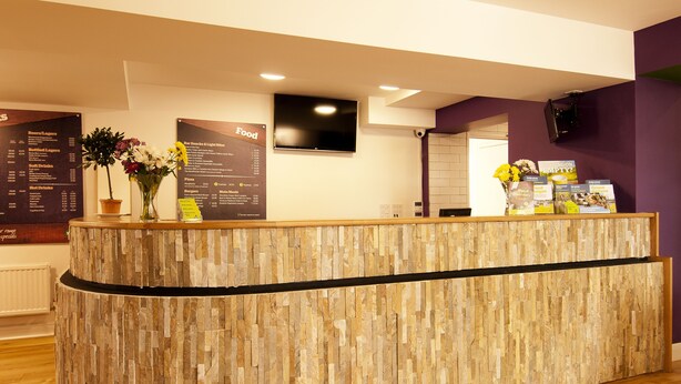 A reception desk with a stone-textured design, a flat-screen TV mounted above it, and a menu board to the left. - YHA York - Hostel (York)