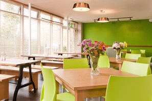 Un restaurant avec des tables en bois et des chaises d’un vert vif, de grandes fenêtres équipées de stores et un mur vert.