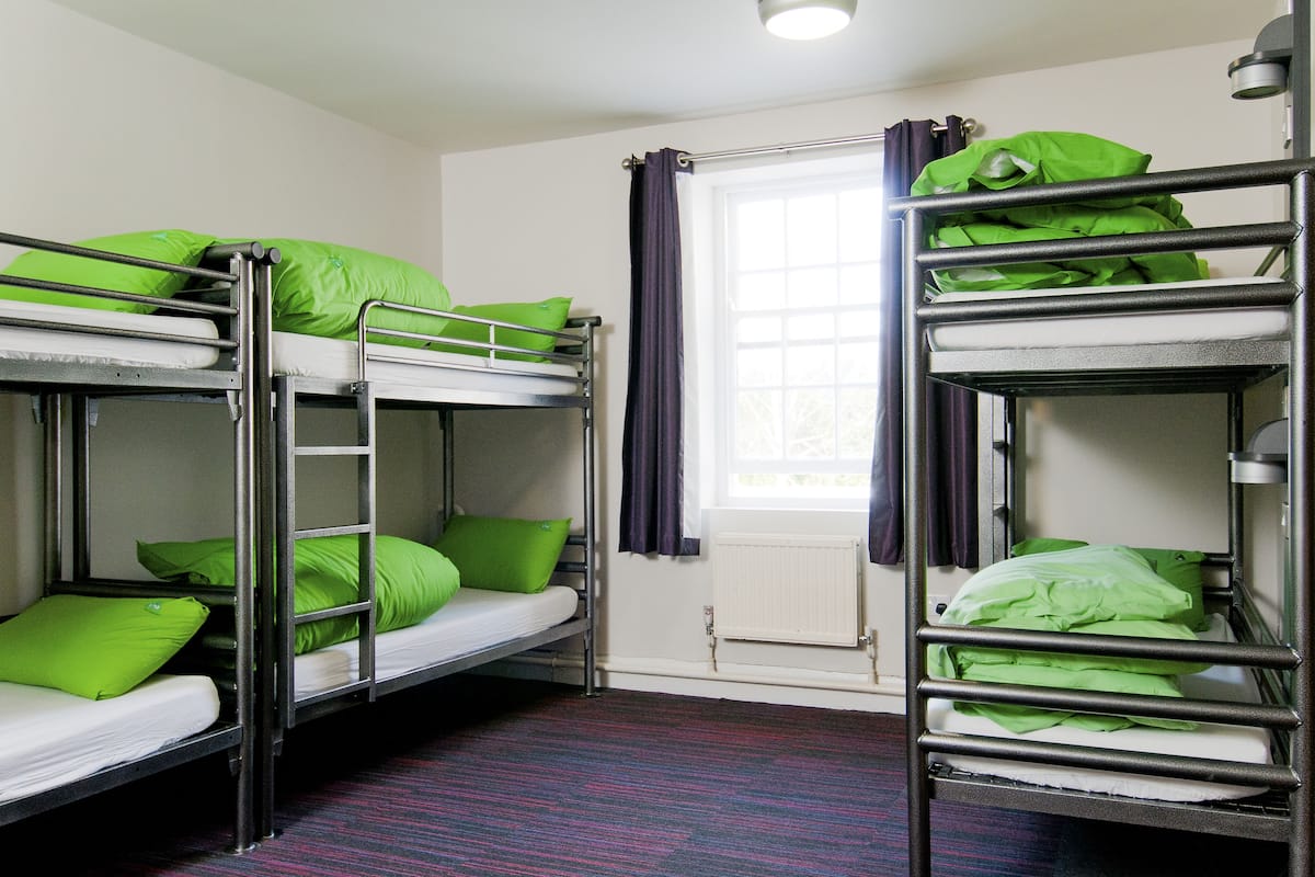 A dormitory room with bunk beds, each with green pillows, a window with curtains, and a radiator.
