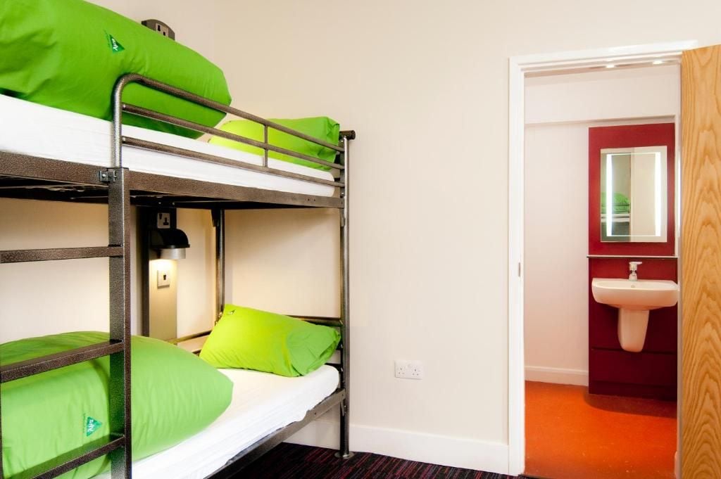 A bunk bed with green bedding, a bedside lamp, and a red-framed mirror in a room with an orange carpet.