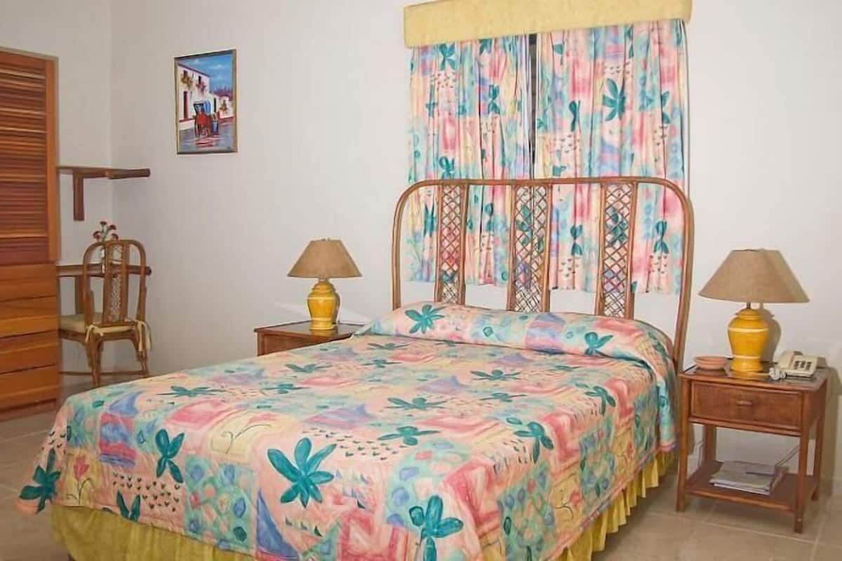 Une chambre à coucher avec un lit, deux tables de chevet avec des lampes, une chaise en bois, une armoire et un tableau accroché au mur.