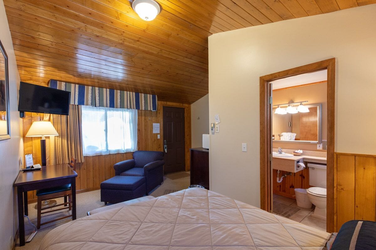 Habitación de hotel con techo de madera, cama, escritorio con lámpara, silla, televisor y baño con espejo y lavabo.