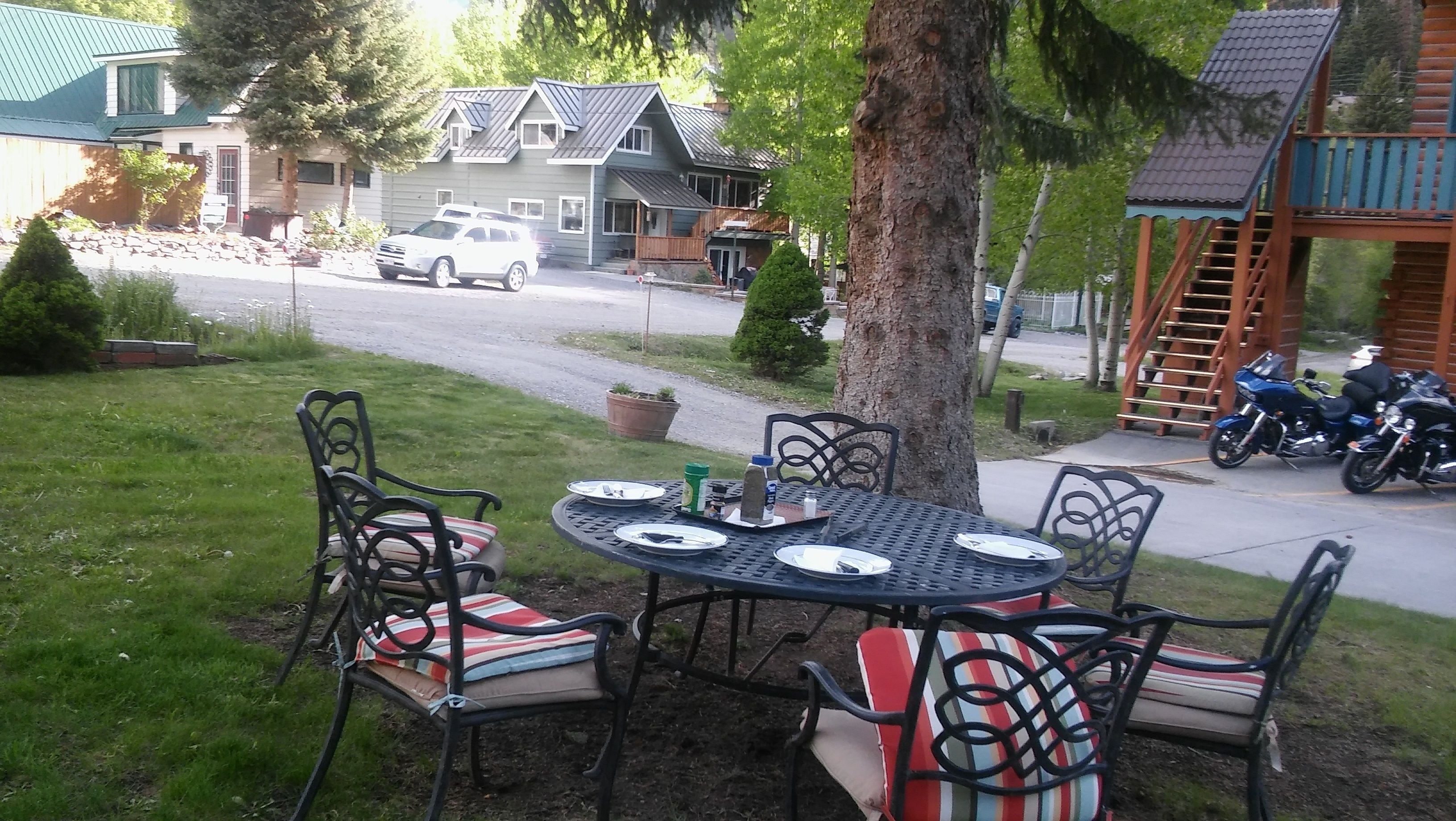 An outdoor dining area with a round table and chairs, a tree, and a view of a street with houses and parked cars.