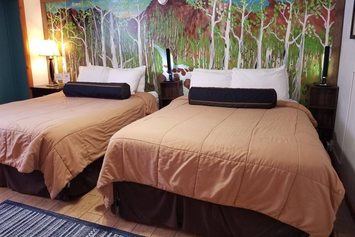 Habitación de hotel con dos camas, ropa de cama color beige, un cuadro de un bosque en la pared, una lámpara y una mesita de noche con un reloj.