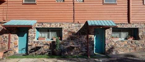Un edificio rústico de paredes de piedra y techos de metal, con tres ventanas pequeñas adornadas con macetas.