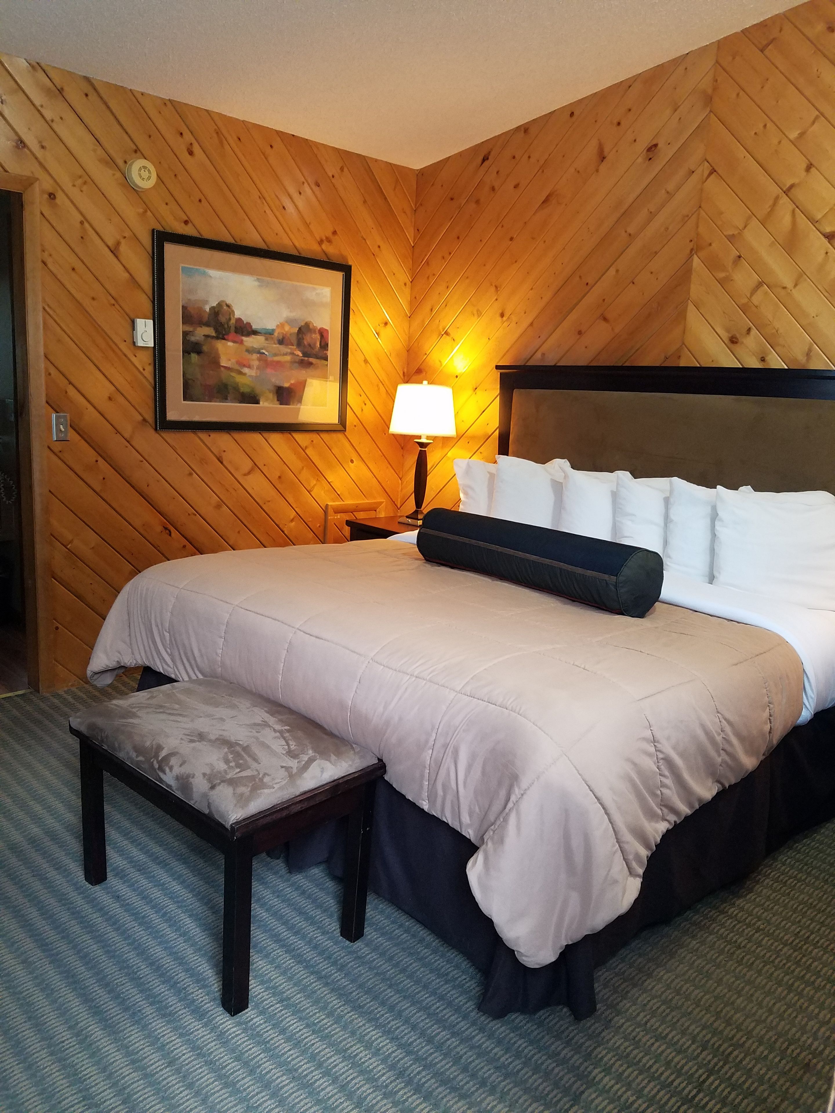 A hotel room with a wooden paneled wall, a bed with white bedding, a nightstand with a lamp, a framed picture on the wall, and a small bench.