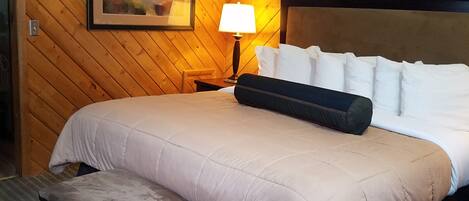 A hotel room with a wooden paneled wall, a bed with white bedding, a nightstand with a lamp, a framed picture on the wall, and a small bench.