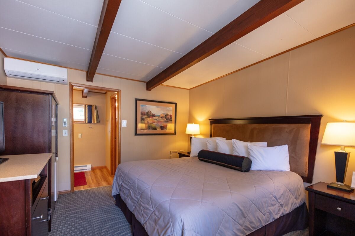 A hotel room with a bed, bedside tables, a desk, and an air conditioning unit.