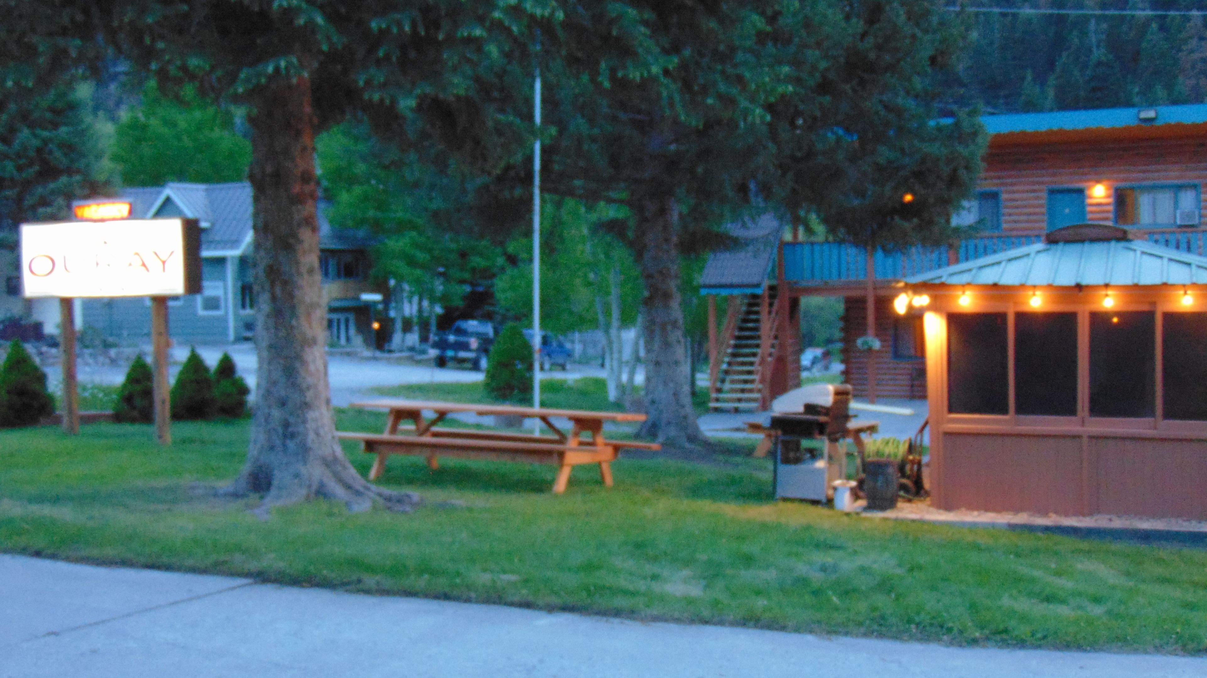 A picnic table and a small gazebo with string lights, surrounded by trees and a building with a sign that reads "OUTWAY".