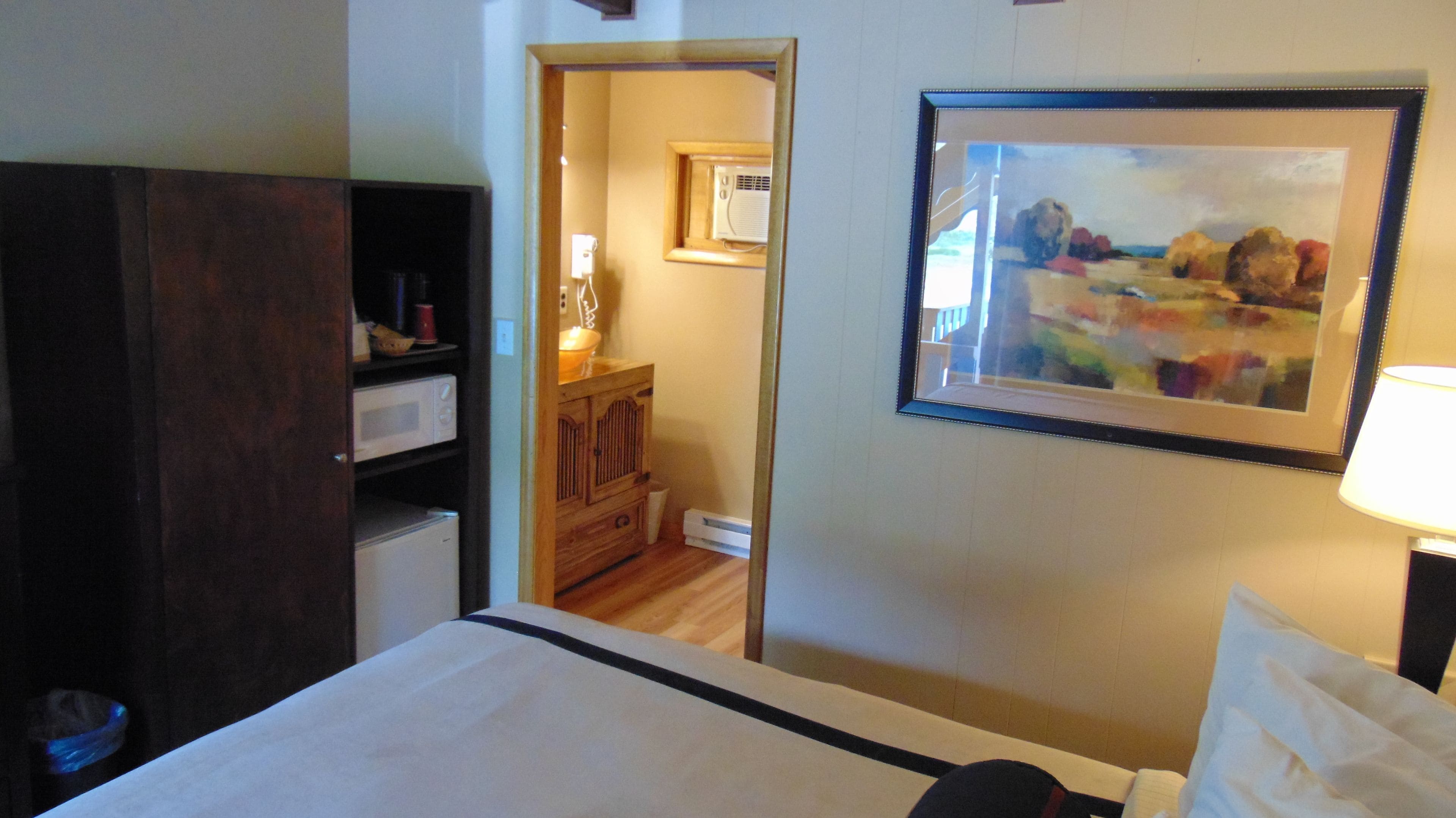 A hotel room with a bed, a wooden wardrobe, a microwave, a small refrigerator, a framed painting, and a lamp.