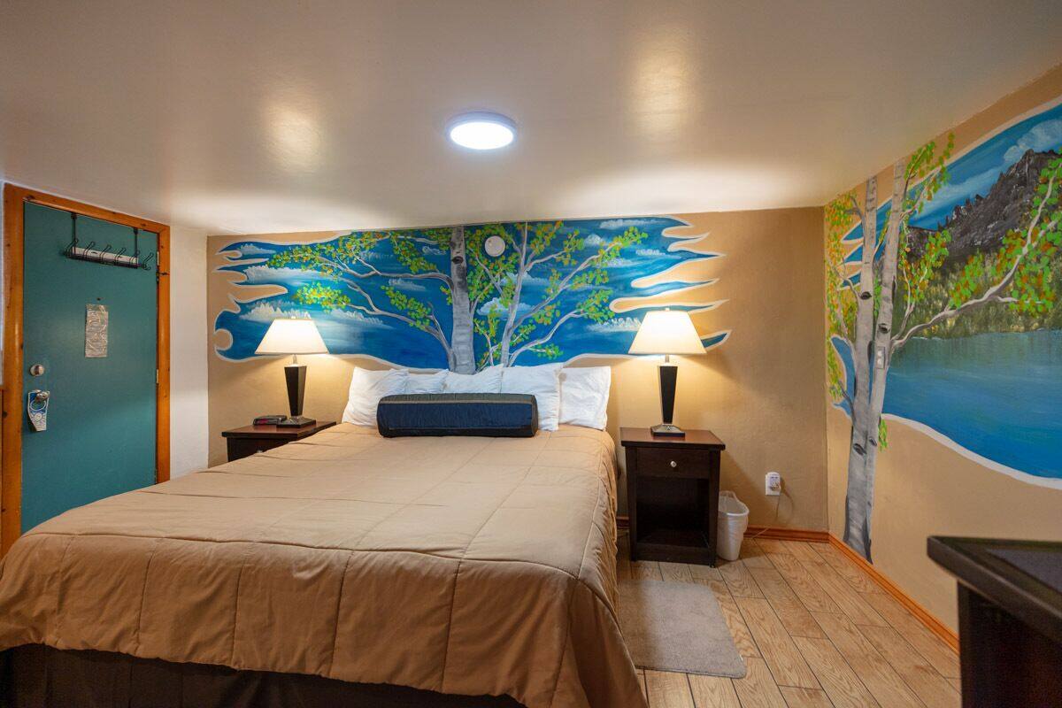Un dormitorio con una cama grande, dos mesitas de noche con lámparas, una mesita auxiliar, una puerta con perchero para toallas y un mural en la pared con un árbol y un paisaje.