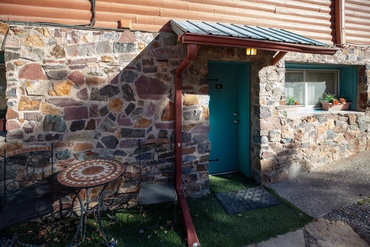 A stone building with a teal door, a small window with plants, and a metal table with a chair outside.