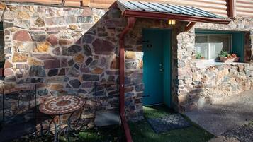 A stone building with a teal door, a small window with plants, and a metal table with a chair outside.