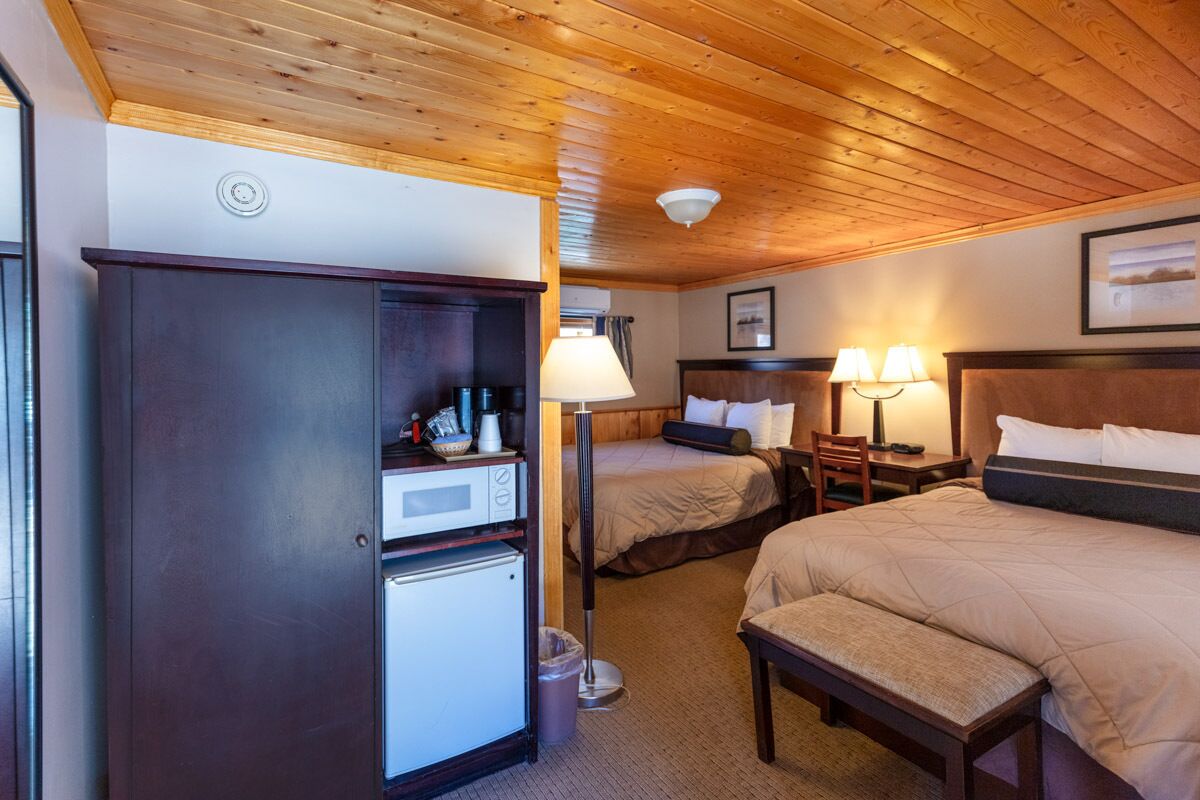 A hotel room with two beds, a desk with a lamp, a wardrobe, and a refrigerator.