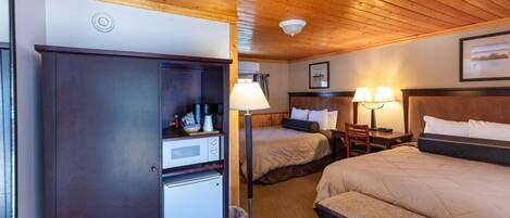 A hotel room with two beds, a desk with a lamp, a wardrobe, and a refrigerator.