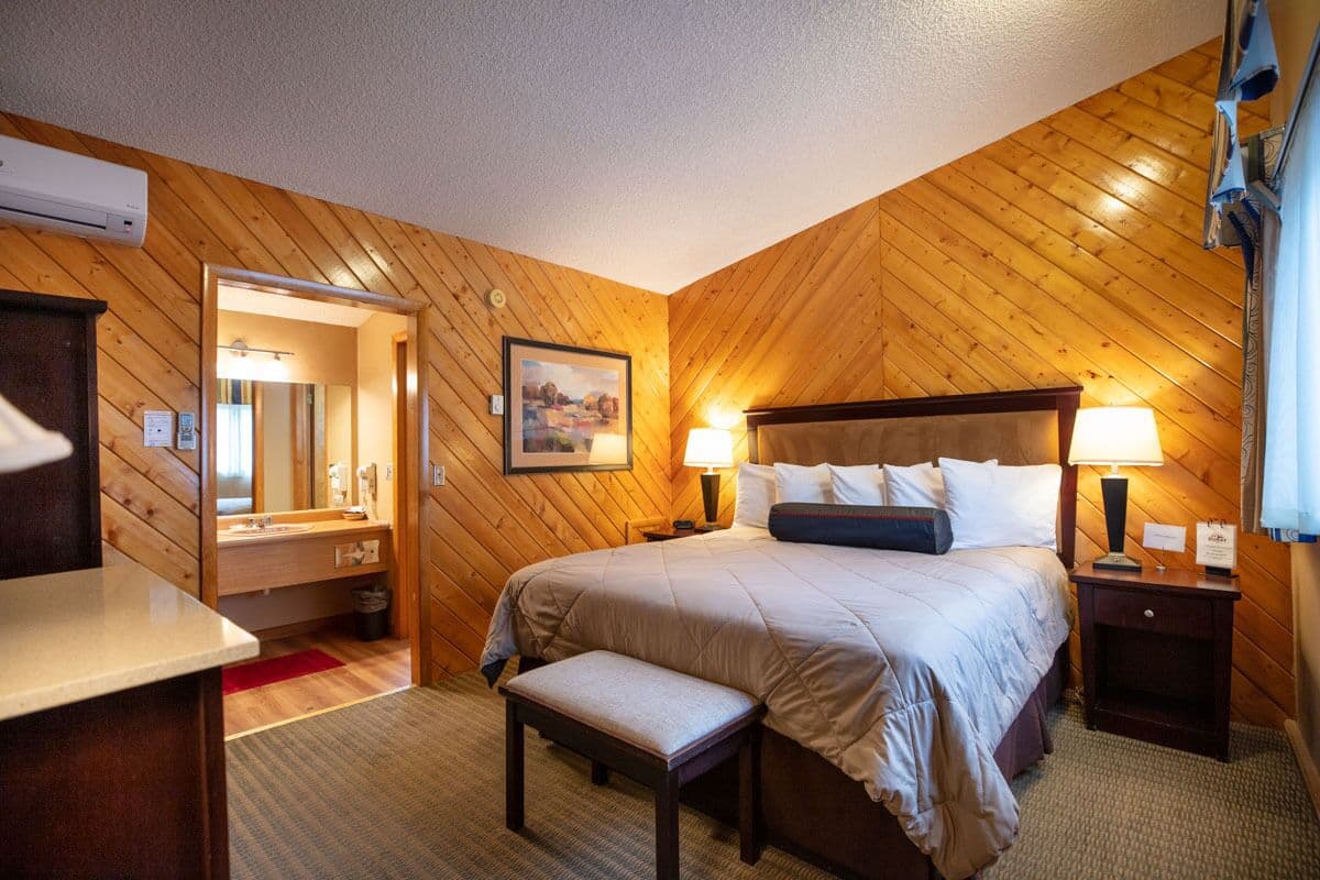 Un dormitorio con techo de madera, una cama con ropa de cama blanca, dos mesitas de noche con lámparas, un banco y un espejo.