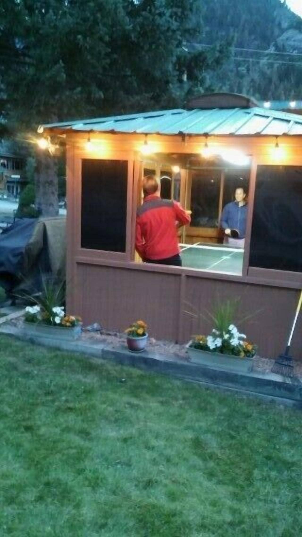 Una pequeña construcción exterior con techo de metal, posiblemente un pabellón o un cobertizo, con una cancha de tenis visible a través de las ventanas.