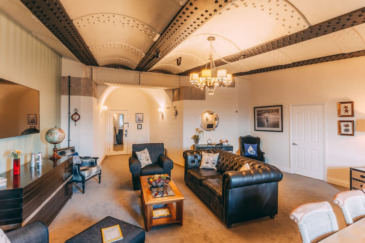 A spacious living room with a leather sofa, armchairs, and a coffee table. A chandelier hangs from the ceiling, and a globe is on a side table.