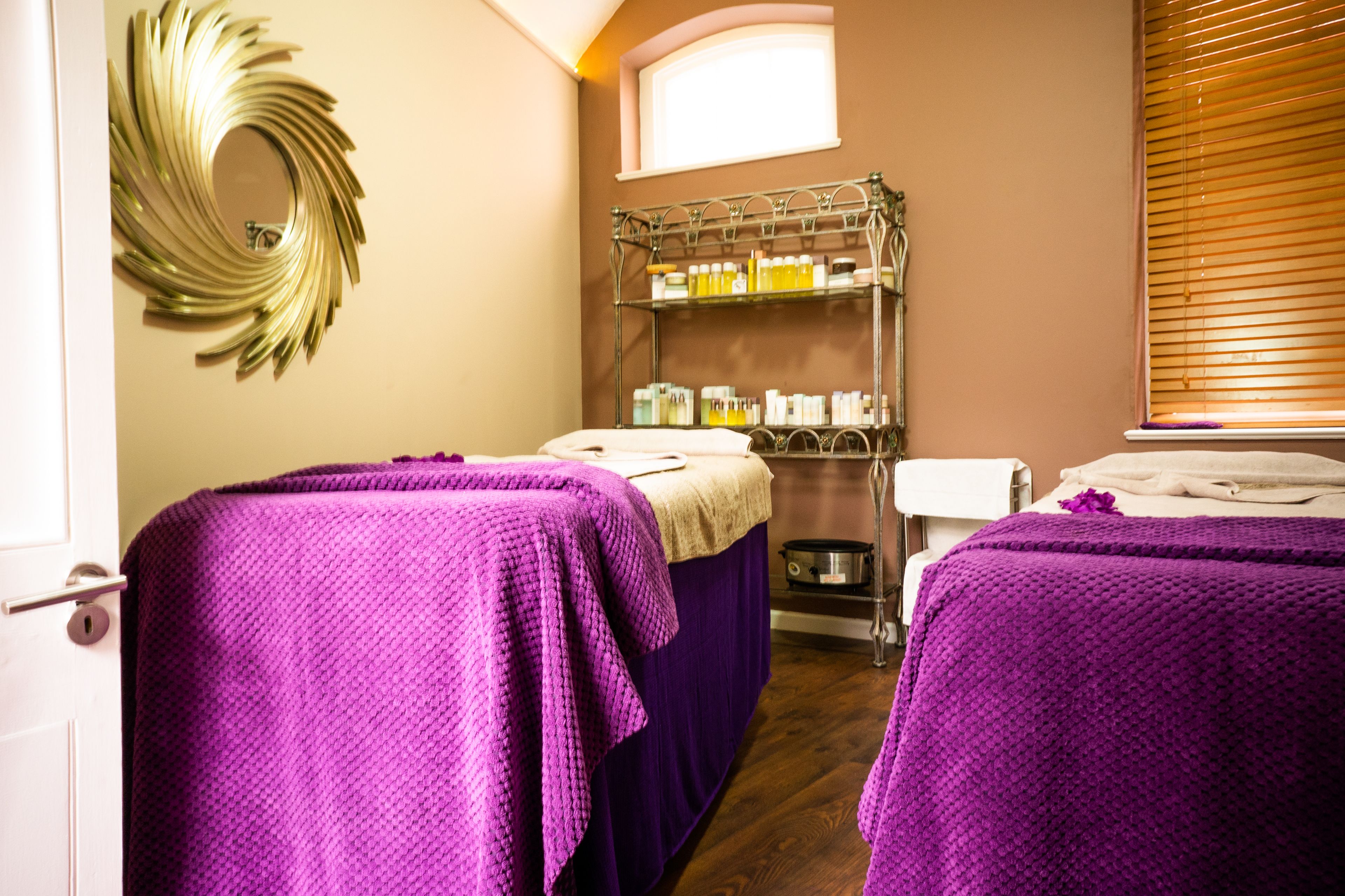 A room with two beds covered in purple blankets, a shelf with toiletries, a mirror, and a window with blinds.