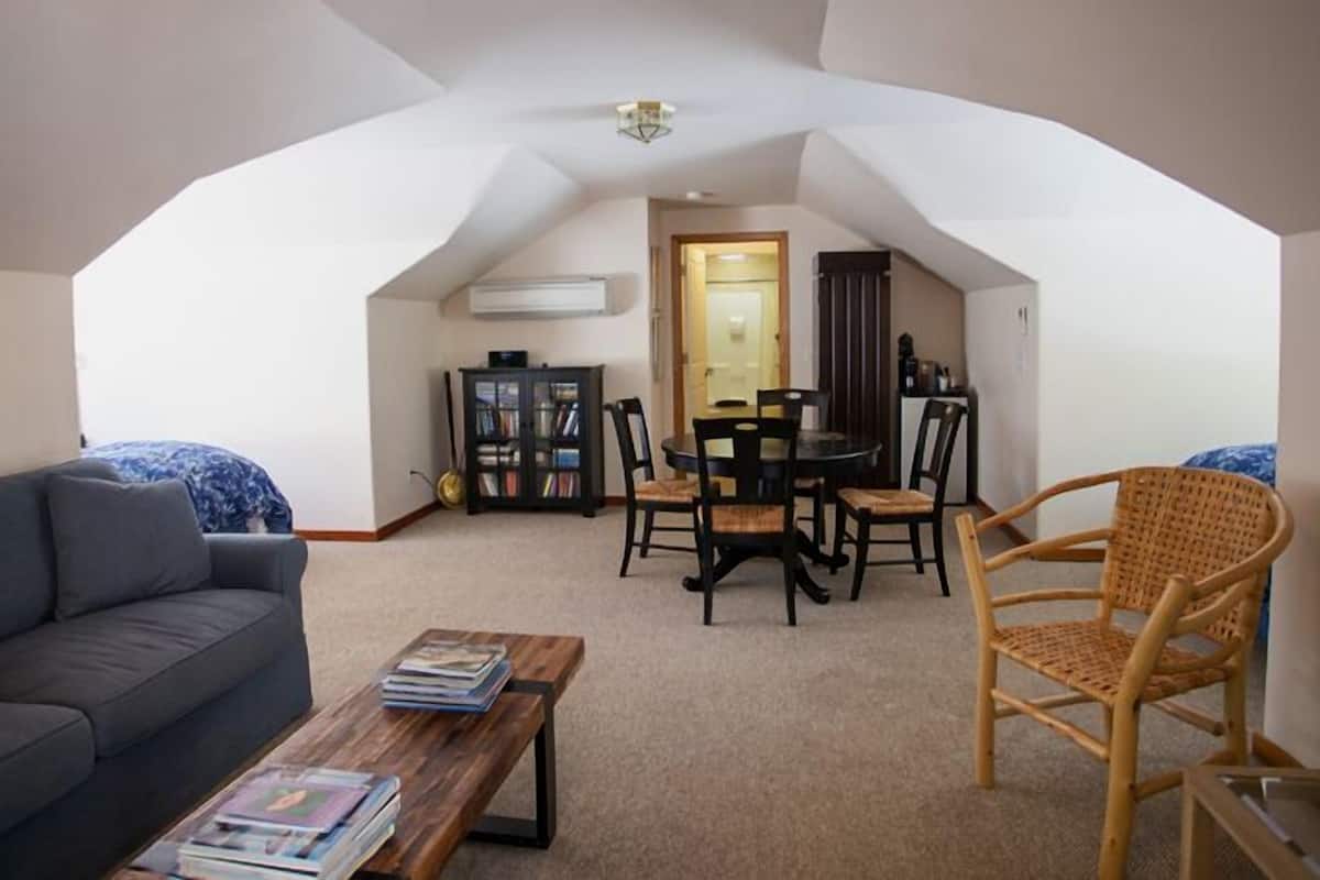 A cozy living room with a sofa, a coffee table, a dining table with chairs, a bookshelf, and a wicker chair.