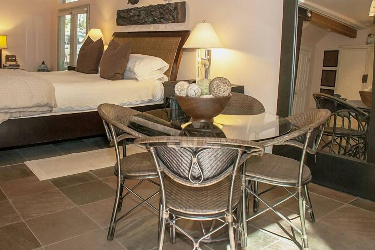 A hotel room with a bed, two bedside tables, a chair, a small table with a bowl, and a view of the outside.