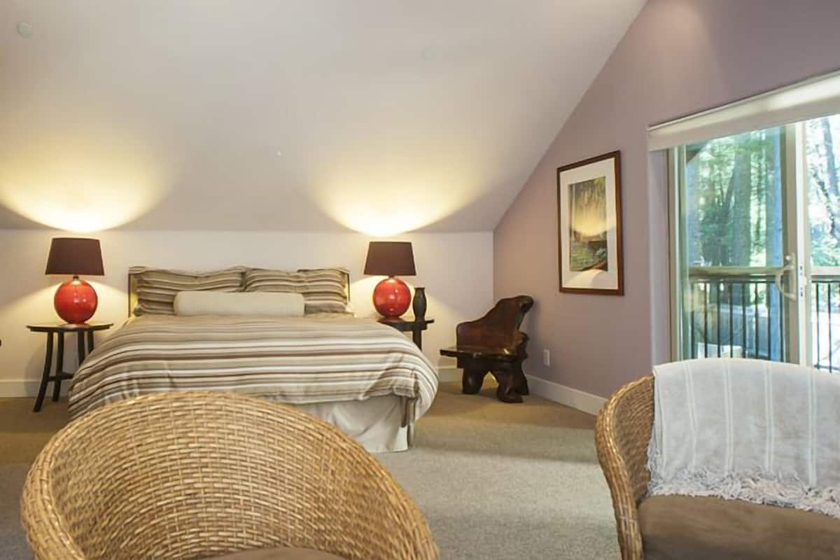 A bedroom with a large bed, two bedside lamps, a wicker chair, and a window with a view of trees.