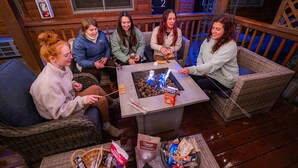 Un groupe de personnes est assis autour d’une table près d’un foyer, sur une terrasse en bois aménagée avec des meubles en osier.