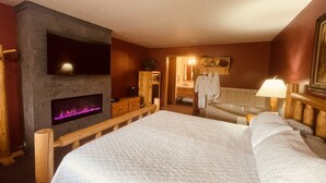 Une chambre d’hôtel avec un grand lit, une cheminée, une tête de lit en bois, une salle de bain avec baignoire et un miroir.