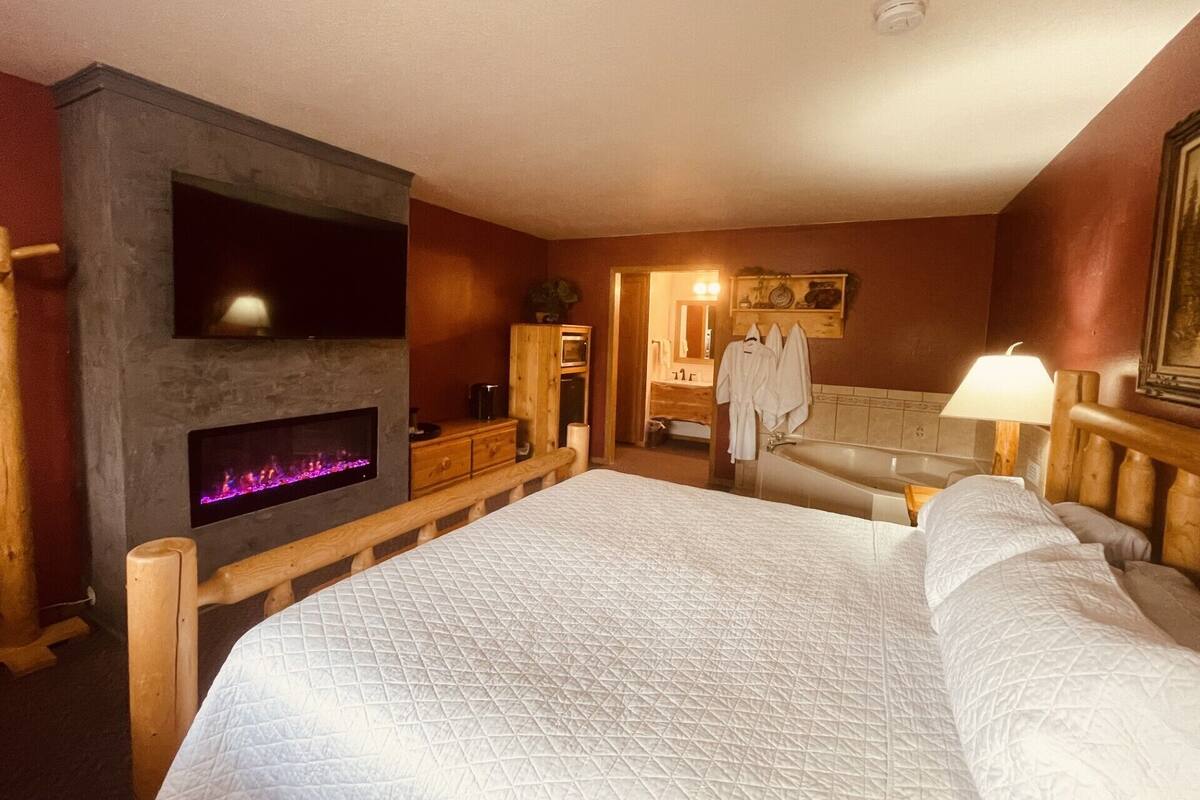 Quarto de hotel com uma cama grande, lareira, cabeceira de madeira, banheiro com banheira e espelho.