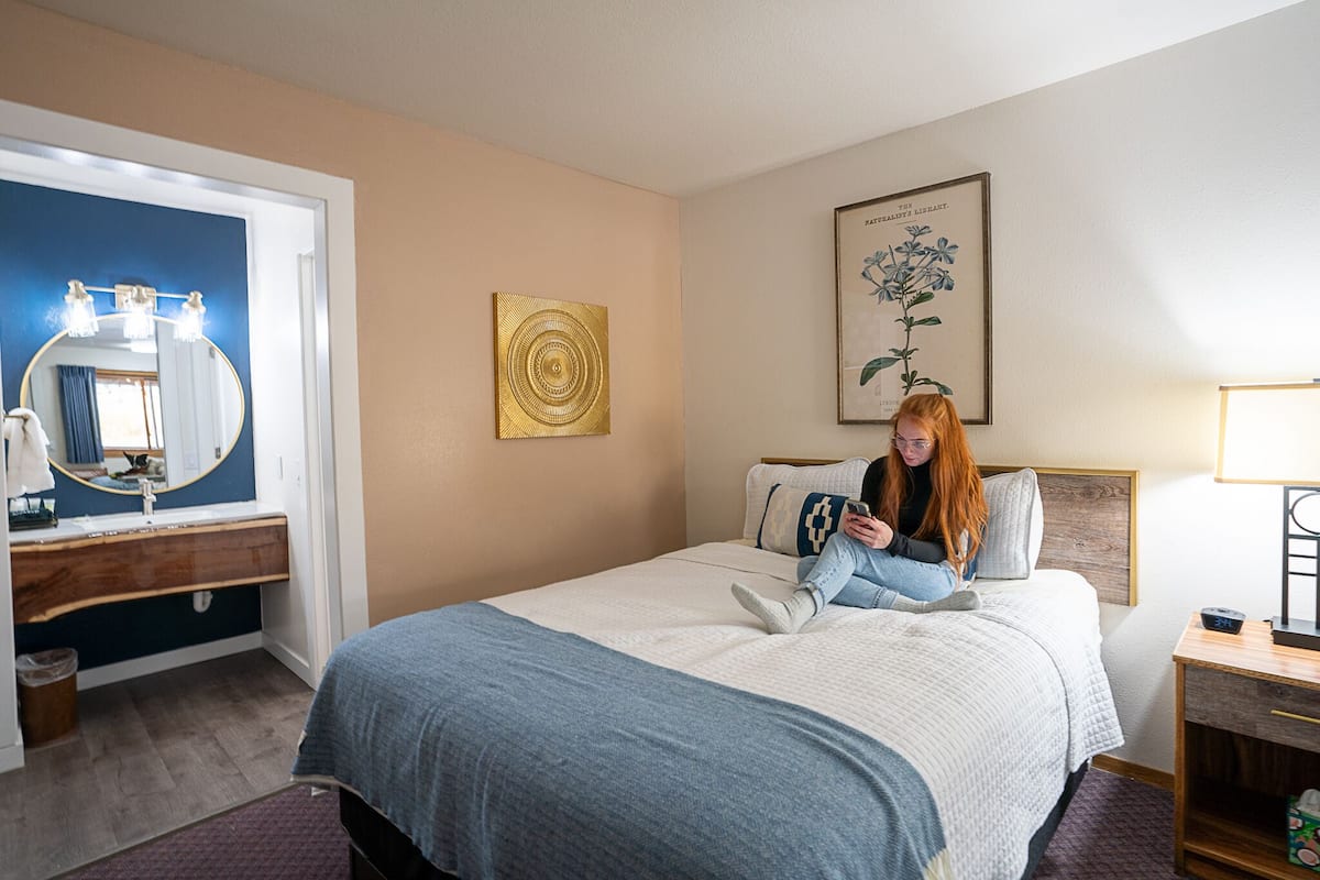 Quarto de hotel com cama, mesa de cabeceira, espelho e uma obra de arte na parede.