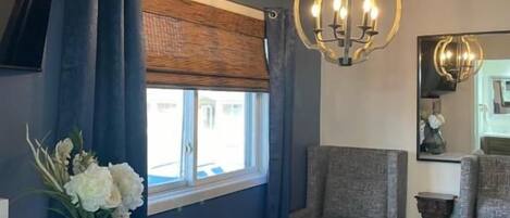Una habitación con una mesa redonda, dos sillas, una lámpara colgante, una ventana con persiana y un florero con flores.
