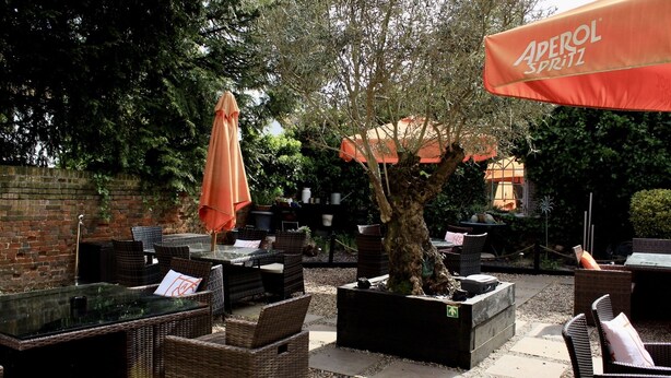 Un espace extérieur aménagé avec des meubles en osier et des parasols orange, entouré de verdure et bordé d’un mur de briques.