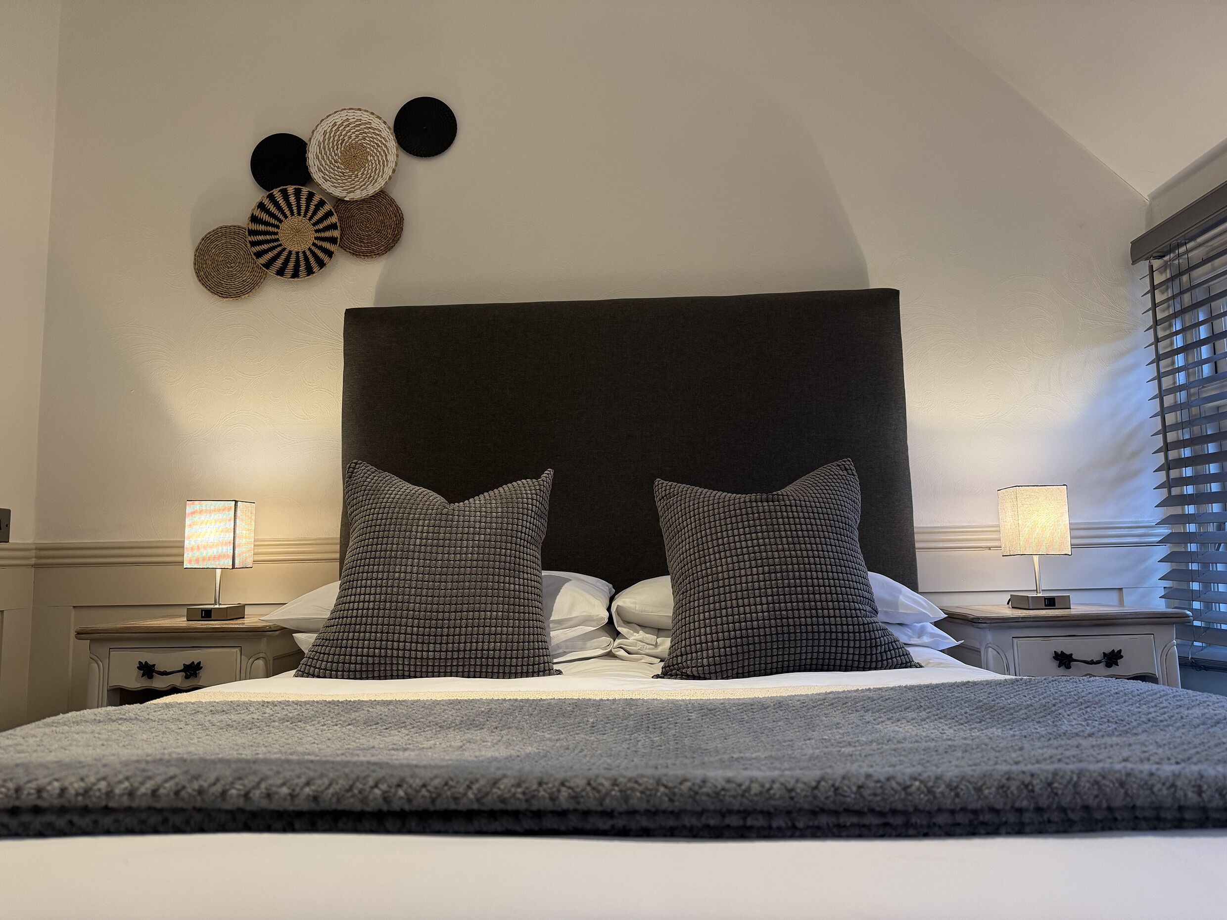 Un lit bien fait, avec deux oreillers, une tête de lit sombre et un mur orné d’objets décoratifs en fibres tressées.