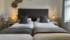 Un lit double avec une tête de lit sombre, deux tables de chevet avec des lampes, un ventilateur fixé au mur et des œuvres d’art décoratives accrochées au mur.
