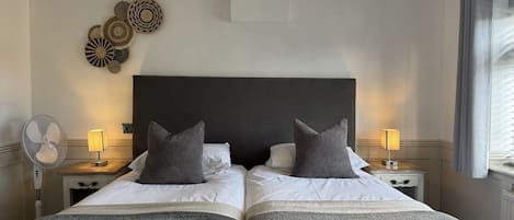 Una cama doble con cabecera oscura, dos mesitas de noche con lámparas, un ventilador de pared y decoración en la pared.