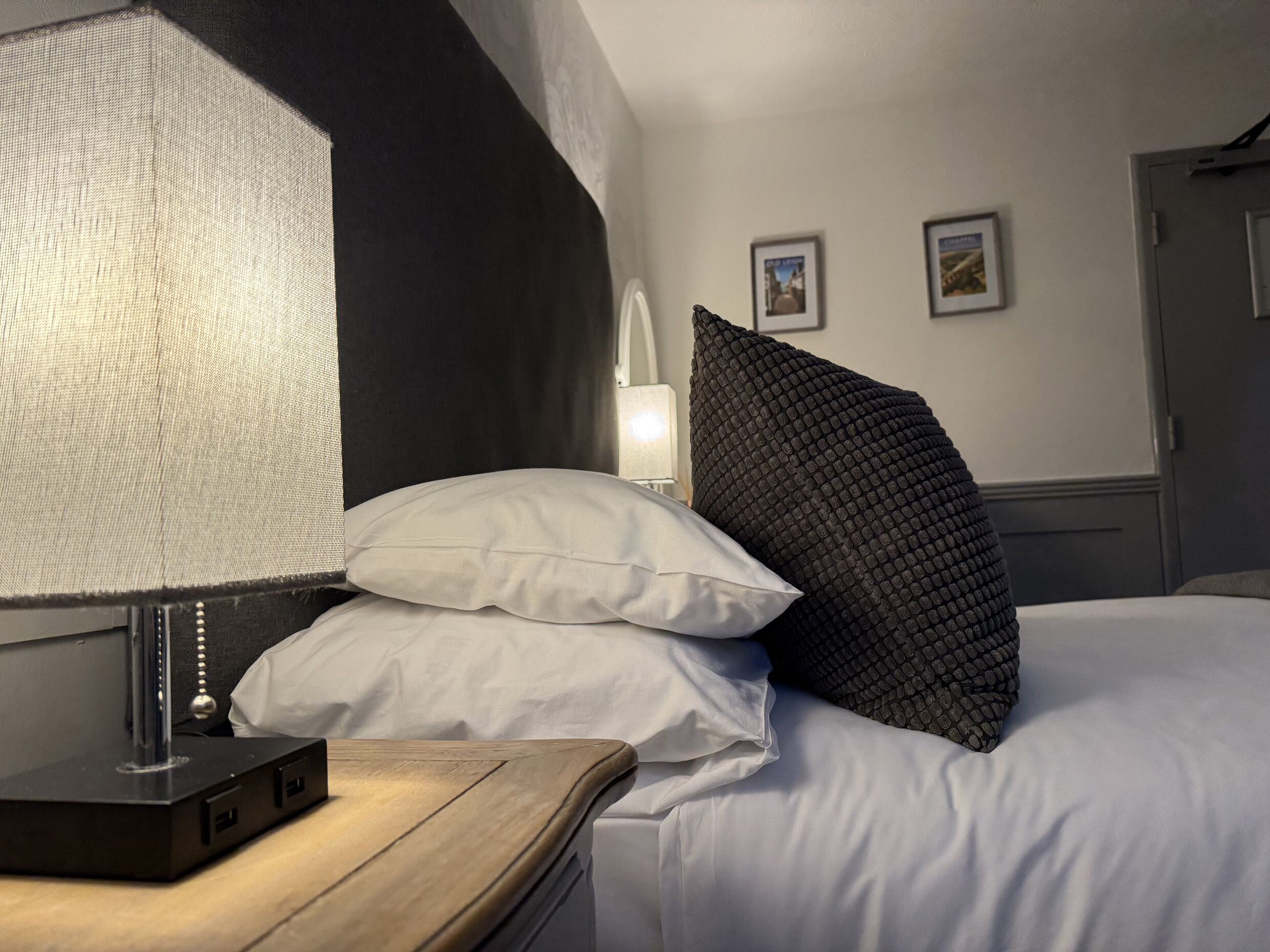 Cama bem arrumada com almofadas brancas e pretas, mesa de cabeceira com abajur e parede com quadros.