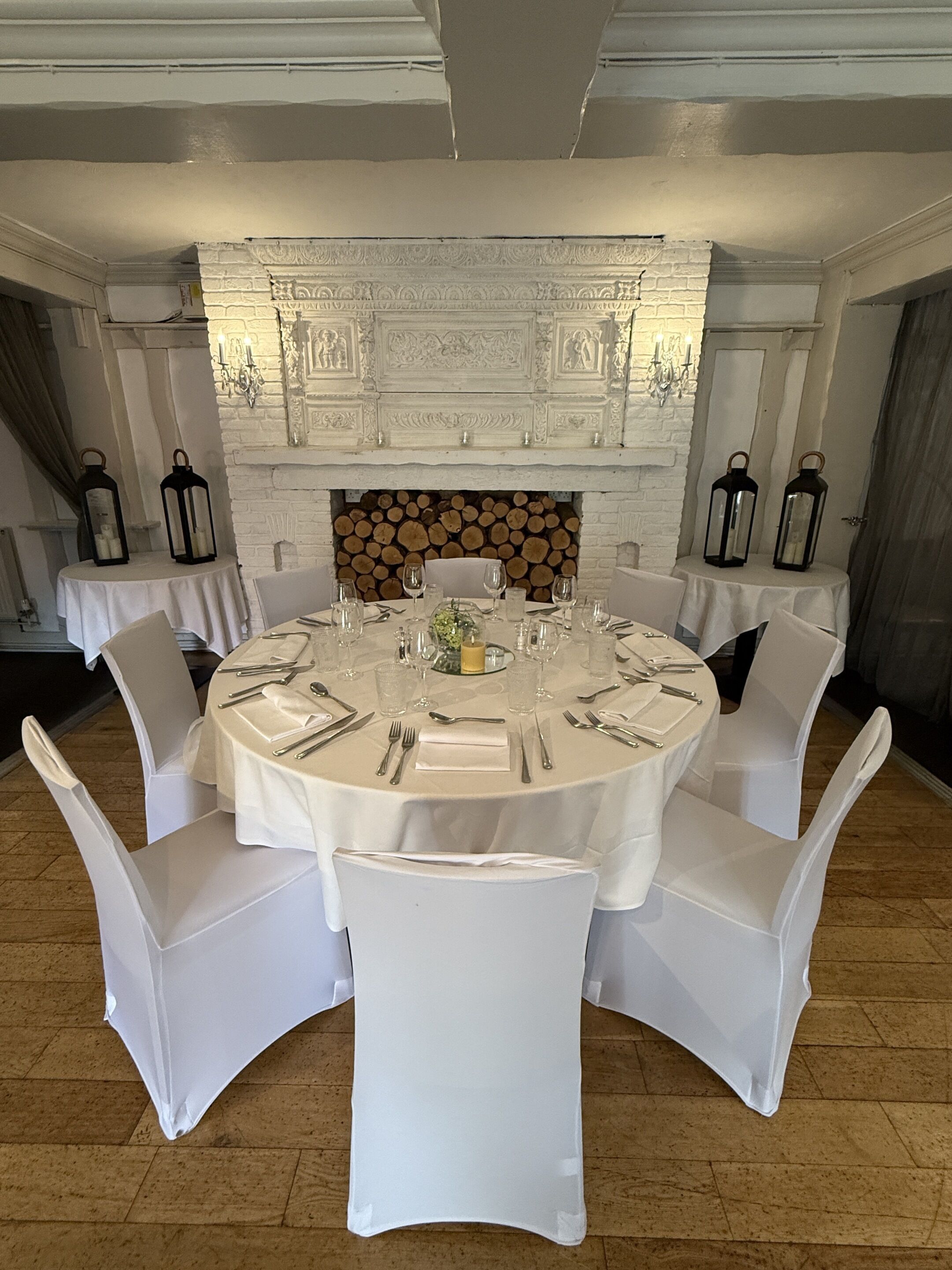 A round dining table set for six with white chairs, a white fireplace with a log fire, and decorative wall sconces.