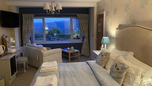 A hotel room with a large bed, a desk with a chair, a small table, a TV, and a window with curtains. - The Gables Bed & Breakfast (Matlock)
