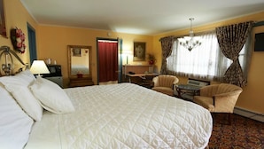 A hotel room with a large bed, a desk, two armchairs, a chandelier, and a window with curtains. - Lantern House Motel (Great Barrington)