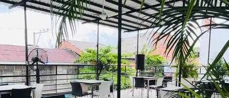 Un área de comedor al aire libre con mesas y sillas, rodeada de grandes plantas verdes y una estructura de techo metálico.