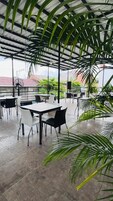 Un área de comedor al aire libre con mesas y sillas, rodeada de grandes plantas verdes y una estructura de techo metálico.
