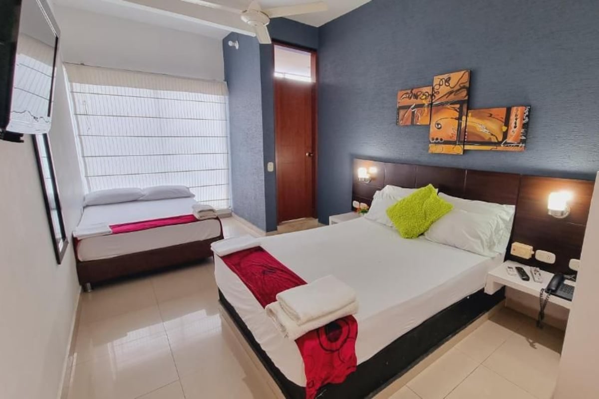A hotel room with a bed, a bedside table, a nightstand, a wall-mounted artwork, and a window with blinds.